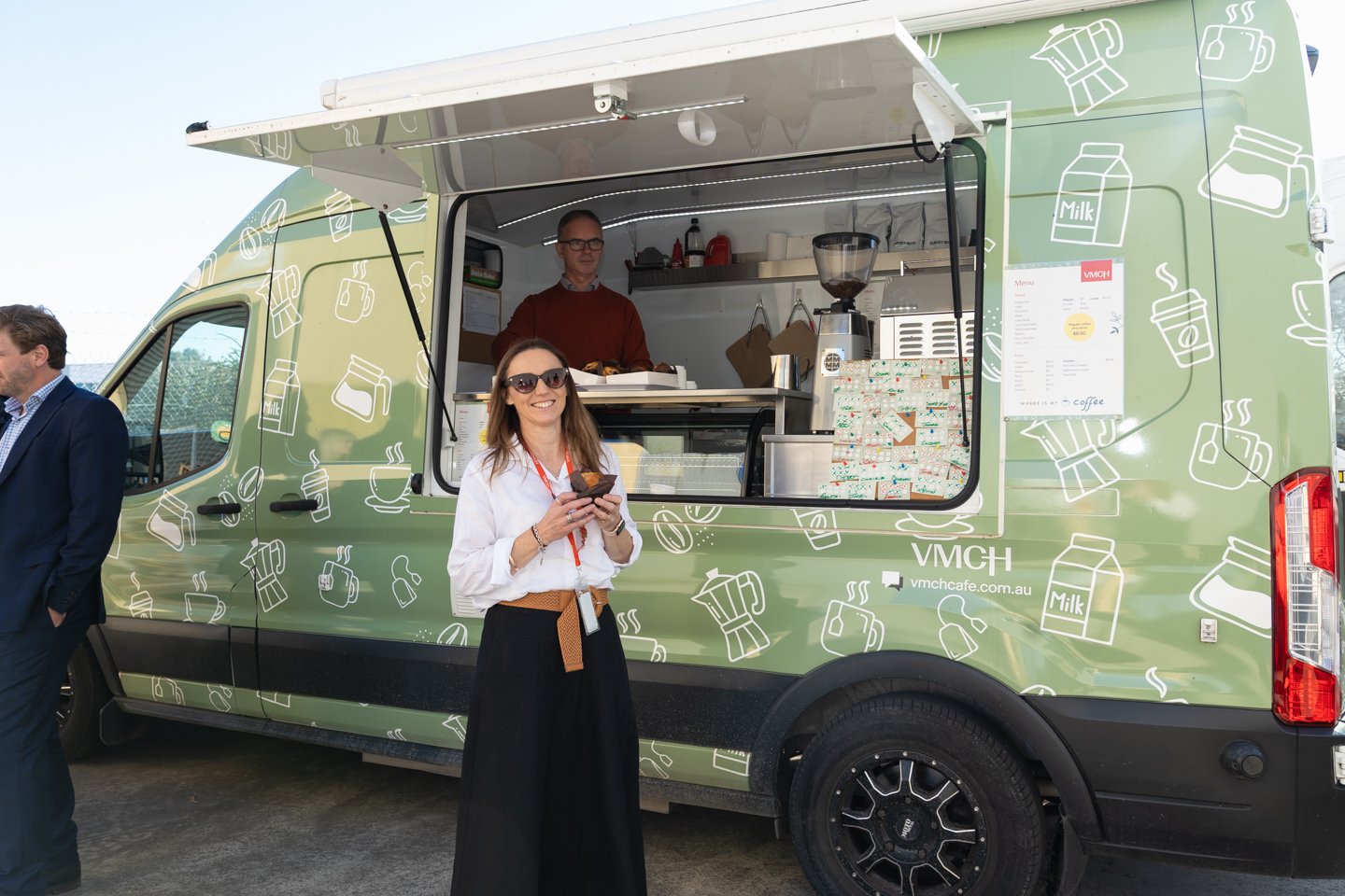 woman wearing white shirt and black skirt standing in front of green coffee van with coffee in hand. Man inside van,
