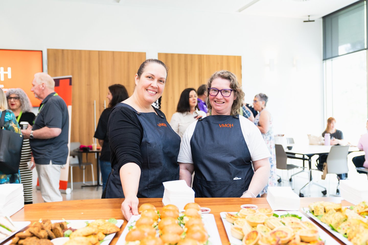 Two women wearing navy apron in fron of food on trays