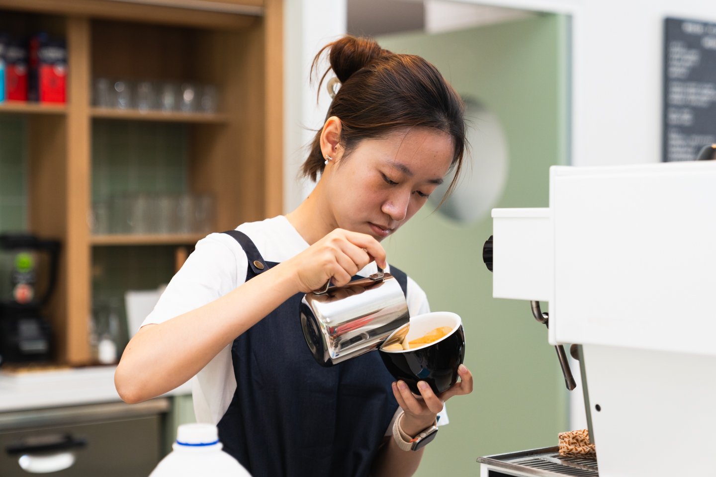 Woman with dark hair wearing white tshirt and navy apron pouring a coffee